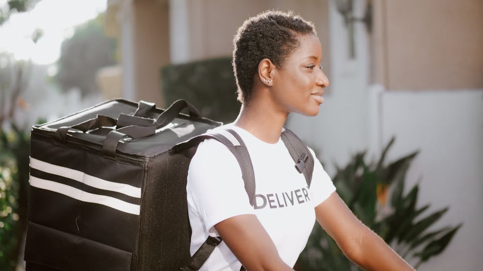 A young woman with dark, curly short hair and medium skin tone, smiling softly in profile as she carries a large black insulated delivery bag with white stripes on her back, wearing a white t-shirt wi