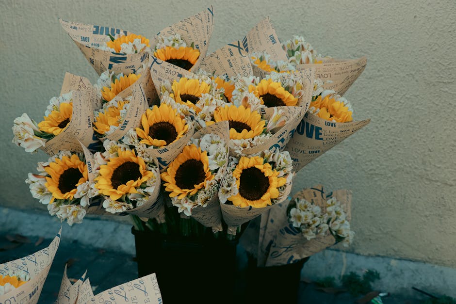 A bouquet of fresh sunflowers with bright yellow petals and dark centers, arranged with small white daisies, all wrapped in brown floral paper with printed words like 'LOVE' and 'LOVE' in blue and bla