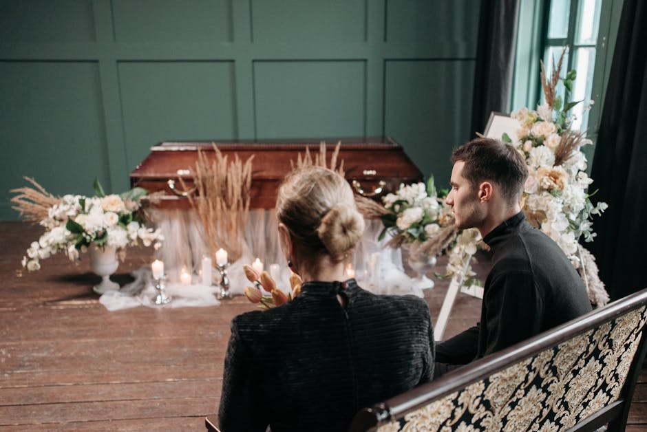A floral tribute setup for a funeral service featuring two mourners seated on a vintage-style black and gold-patterned bench, facing a wooden casket surrounded by white and cream-colored flowers, incl