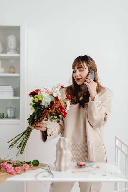 A woman with long brown hair, wearing a beige sweater, is holding a large, colorful bouquet of fresh flowers including red, white, pink, and green blooms, in one hand while talking on a mobile phone w