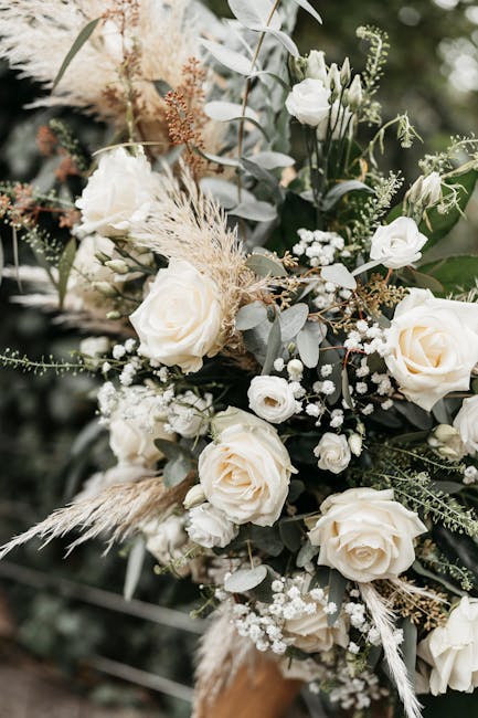 A close-up of an elegant floral arrangement featuring large, creamy white roses with gently unfurling petals, complemented by small white lisianthus and delicate baby's breath. The bouquet incorporate