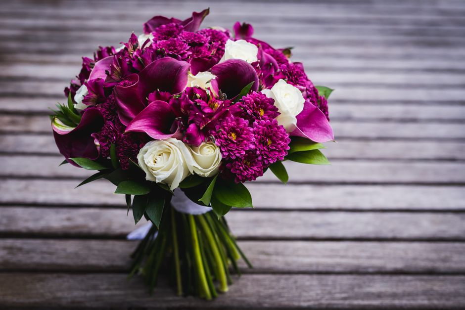 A bouquet of fresh floral arrangement featuring deep purple calla lilies, bright pink chrysanthemums, and white roses, complemented by green foliage and wrapped in white ribbon, resting on a weathered
