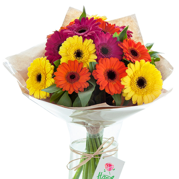 Centered in this evocative scene is a lavish arrangement of Gerbera daisies, their luminous hues catching the light in a way that enlivens any Shacklewell living space. The bouquet displays an expert combination of large, sun-bright yellow, zesty orange, and jewel-toned magenta blooms, each radiating from velvety, near-black centers that bring dramatic depth to the floral array. Interlaced with lush green foliage, the daisies appear anchored in a natural tapestry reflective of the area's hidden gardens. The upper portion of the bouquet is enveloped in gently crumpled light brown kraft paper, offering a rustic counterpoint to the modern clarity of the cellophane wrapping, which rises protectively around the blossoms. The sturdy stems gleam through the transparent, slightly hourglass-shaped vase-a nod to fresh, flowing water drawn just that morning. Around the stems, a soft, natural twine is tied in a simple bow, giving an artisanal finish reminiscent of Flowers Shacklewell's signature touch. Suspended from the bow, a white tag delicately features a sketched red rose, its partially visible script a secret waiting to be discovered. The arrangement is equally fit for celebratory gatherings at local Shacklewell cafés, thoughtful housewarmings, or simply to welcome warm sunlight streaming into a community home. This floral display is a vibrant ode to Shacklewell's spirit, perfect for same-day delivery or sharing a radiant gesture that lingers well beyond the first glance.